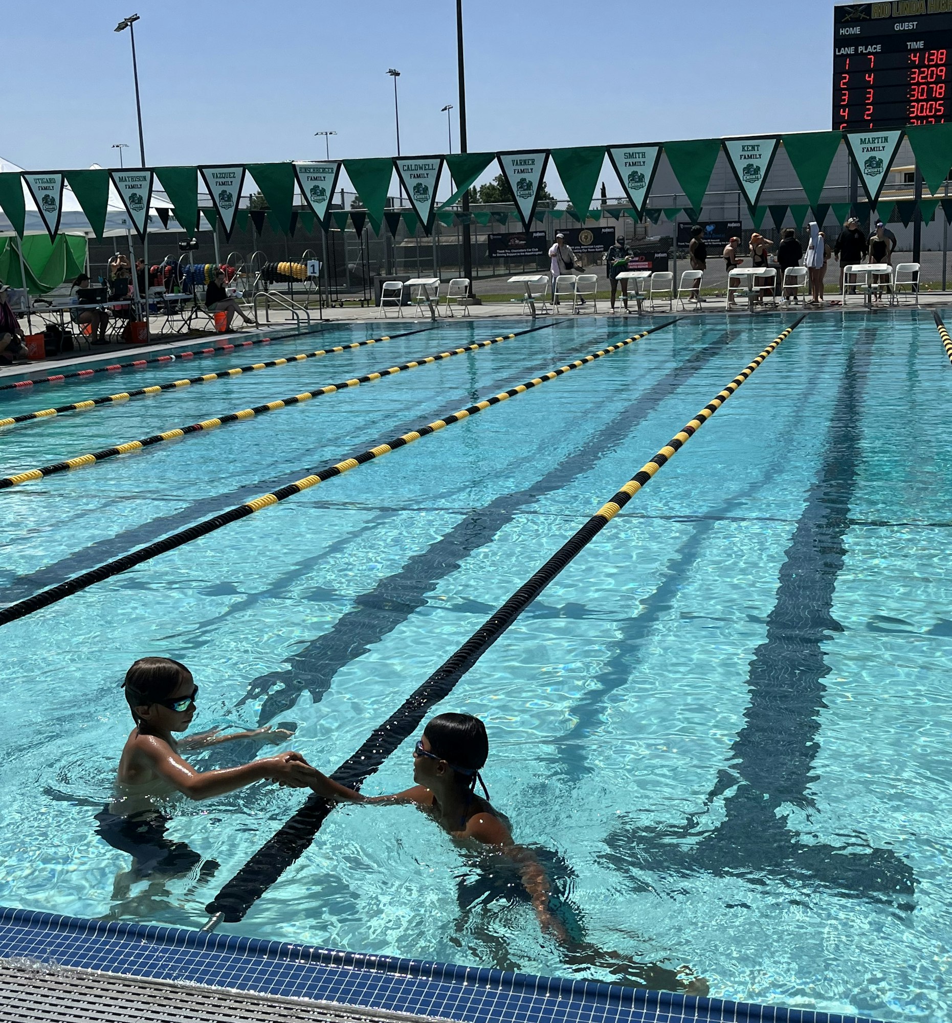 Two swimmers in a pool shake hands after a race; timing board and flags visible in the background.