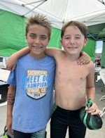 Two young boys standing together, smiling under a tent; one is shirtless.