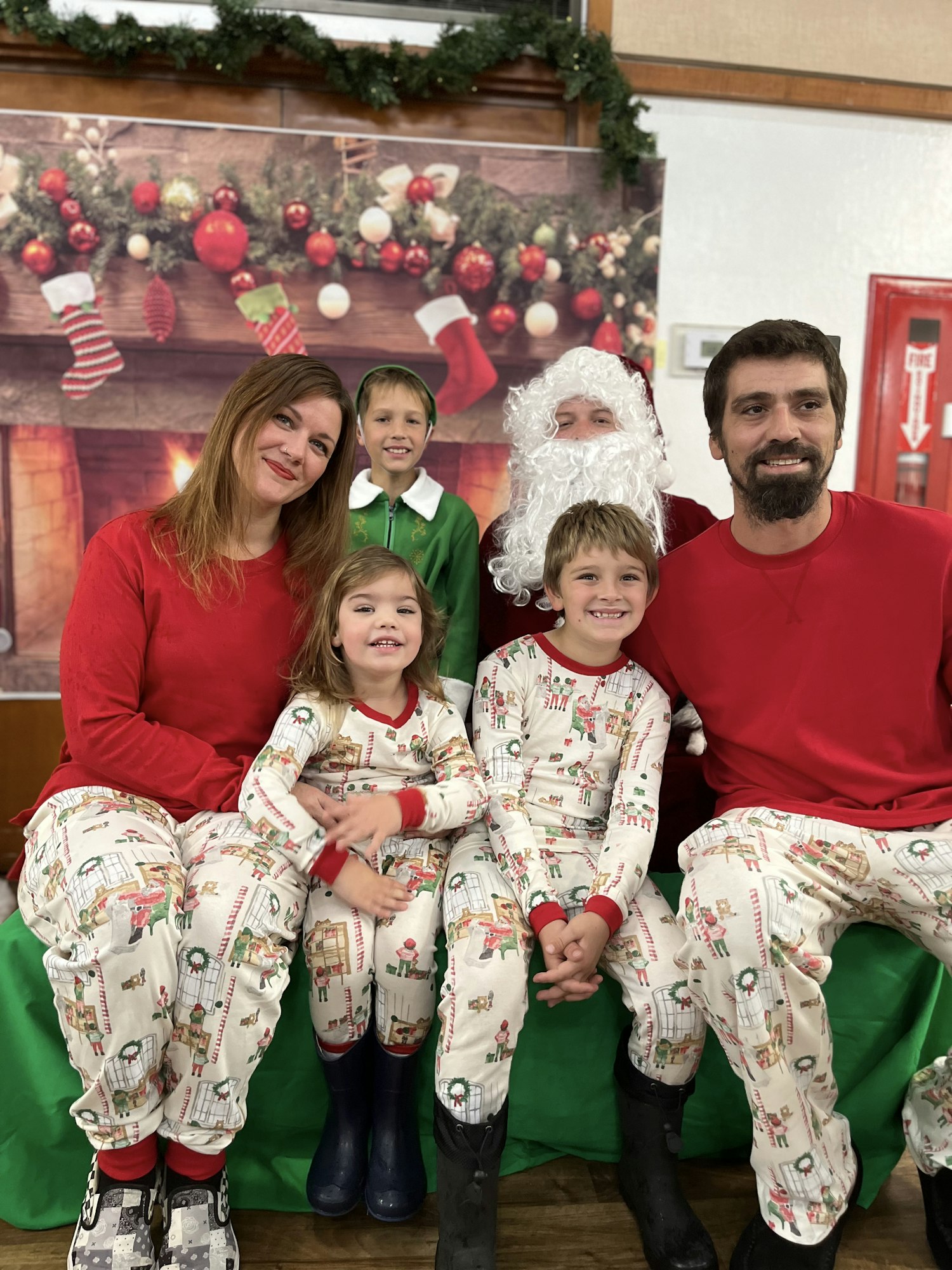 A family in matching holiday pajamas poses with a person dressed as Santa in front of a festive fireplace backdrop.