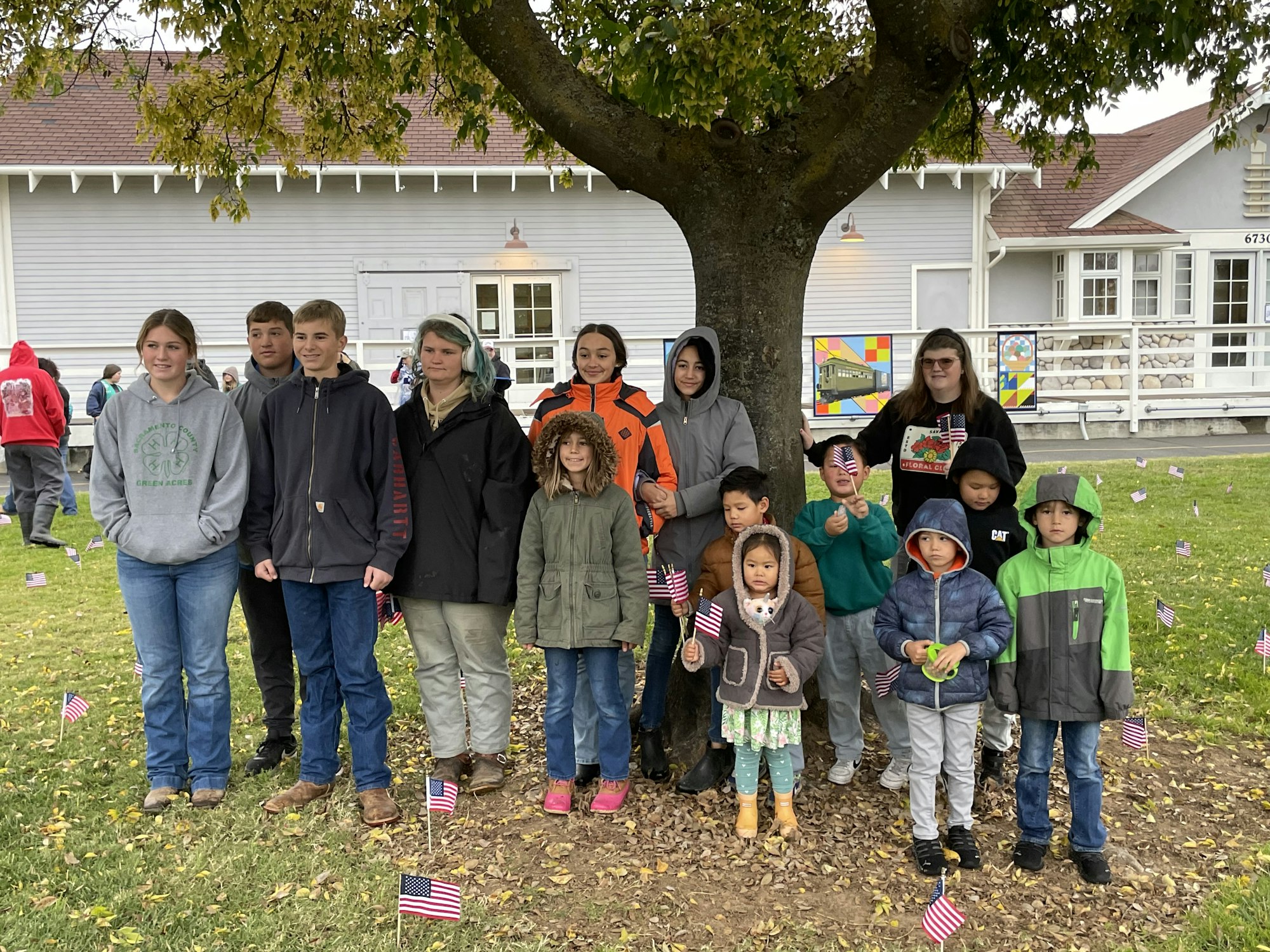 A group of people, both adults and children, stand under a tree holding small American flags, with a building in the background.