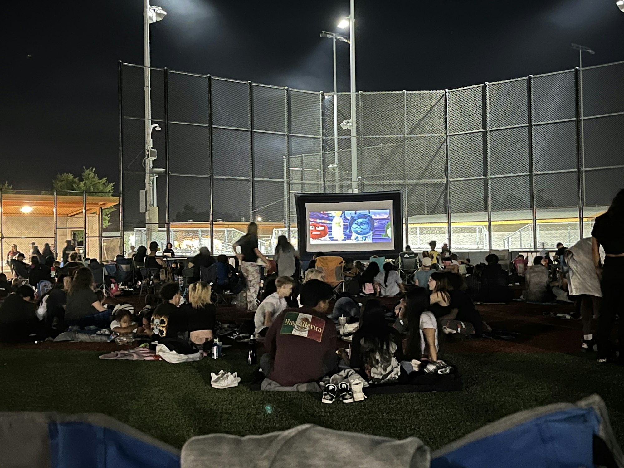 Outdoor movie night with people sitting on grass, watching a screen, surrounded by sports field fencing.