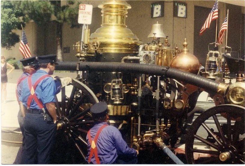 The image shows a vintage steam engine, with men in uniform inspecting it, flags in the background, and a no-parking sign visible.
