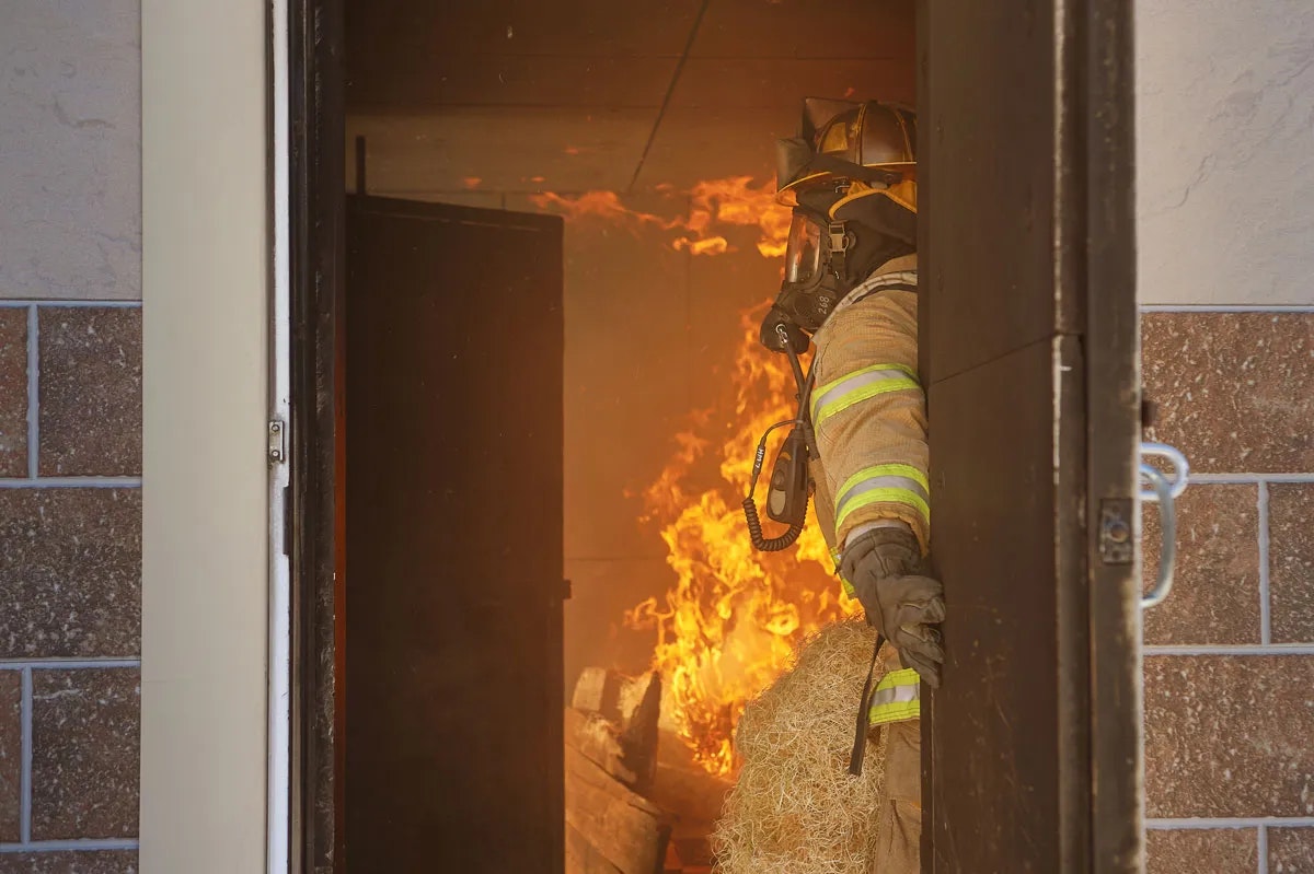 A firefighter stands near a door, facing a blazing fire inside a building, equipped with gear and a breathing apparatus.
