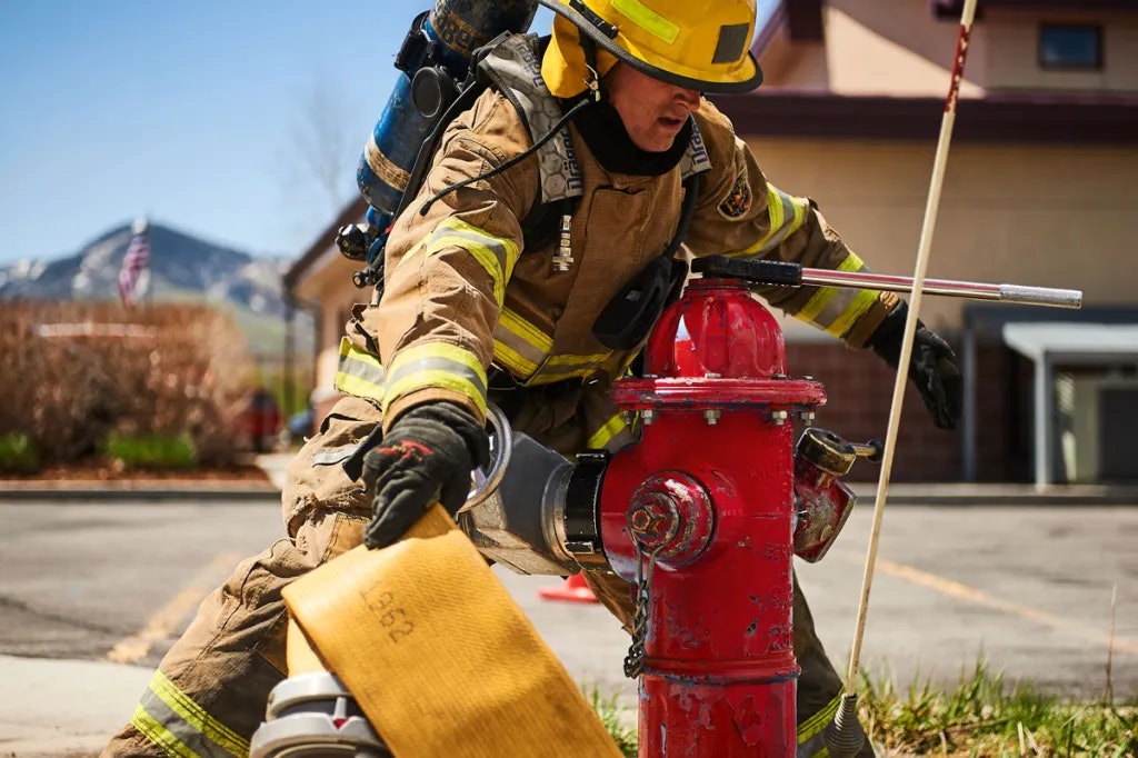 A firefighter in gear operates a fire hydrant, preparing to connect hoses, with a mountainous backdrop and bright day.