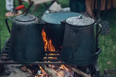 The image shows two black metal containers over a campfire, surrounded by greenery, suggesting outdoor cooking or camping.