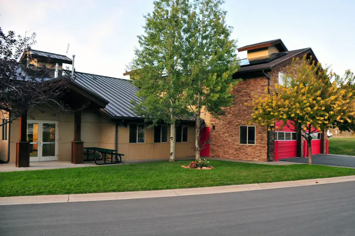 A modern building with a combination of stone and stucco, surrounded by greenery and trees, featuring a red garage door.