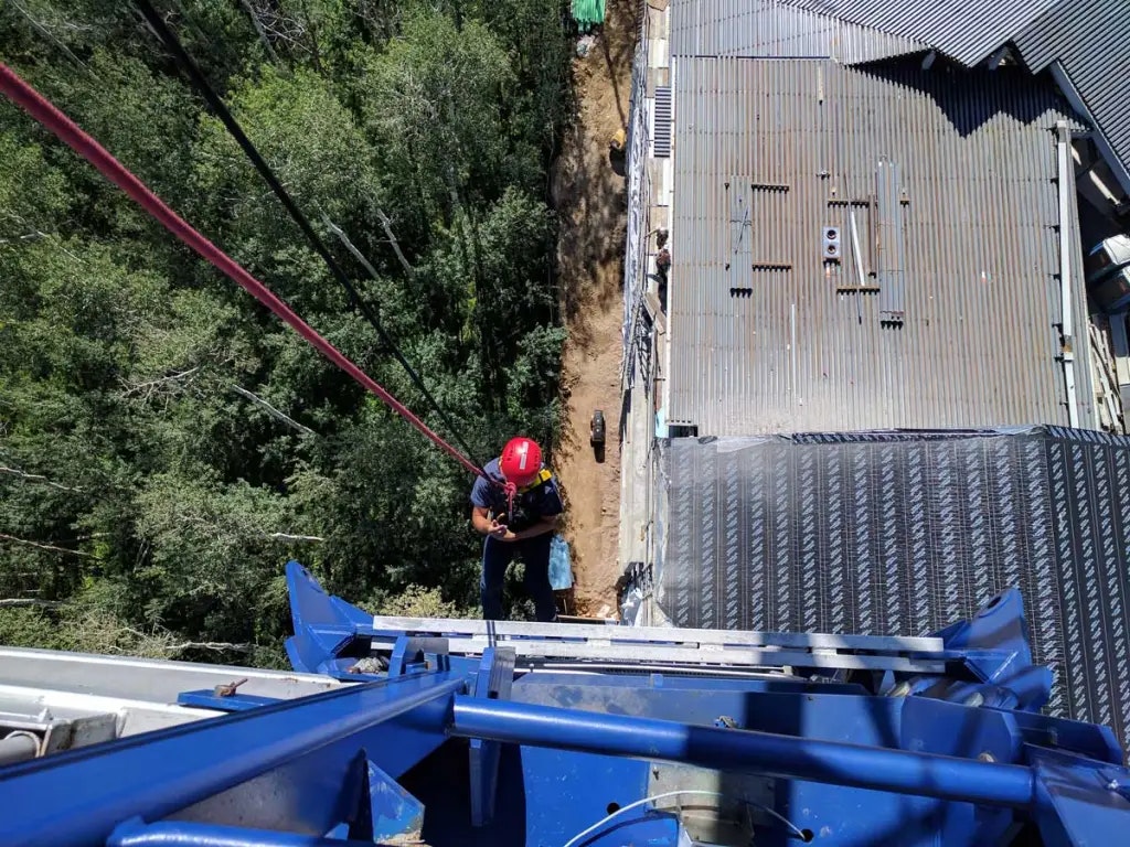 A person in a helmet is rappelling down a tall structure, with trees and a building visible below.