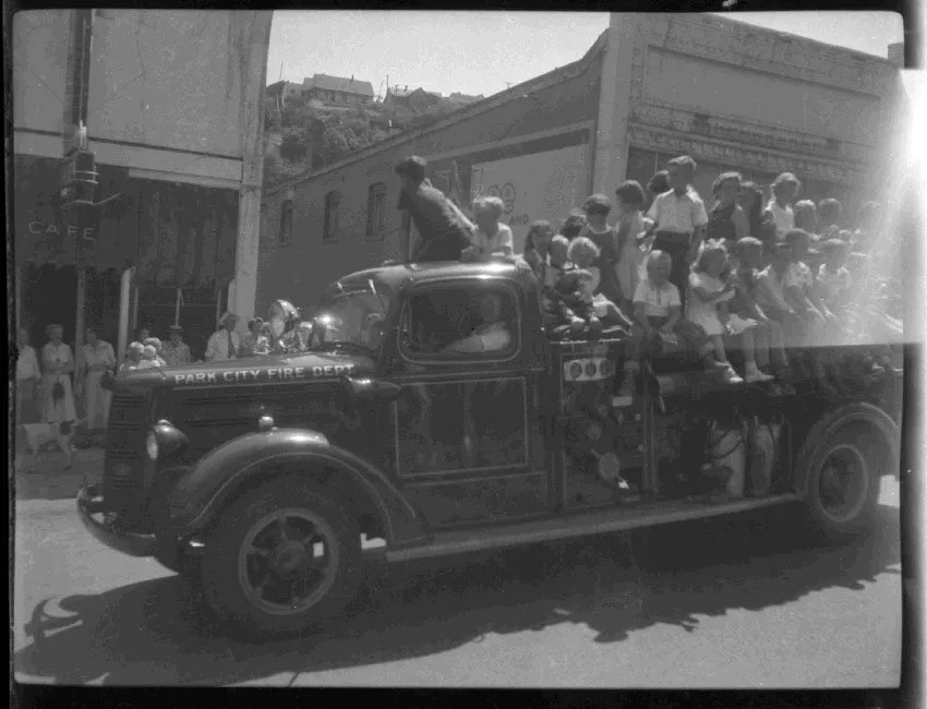 A vintage fire truck with children sitting on it drives through a street, while onlookers watch from the sidewalk.