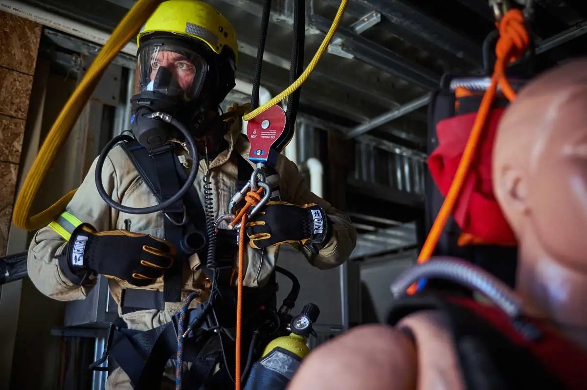 A person in a safety helmet and gear is preparing for a rescue operation, handling a harness and ropes. A mannequin is nearby.
