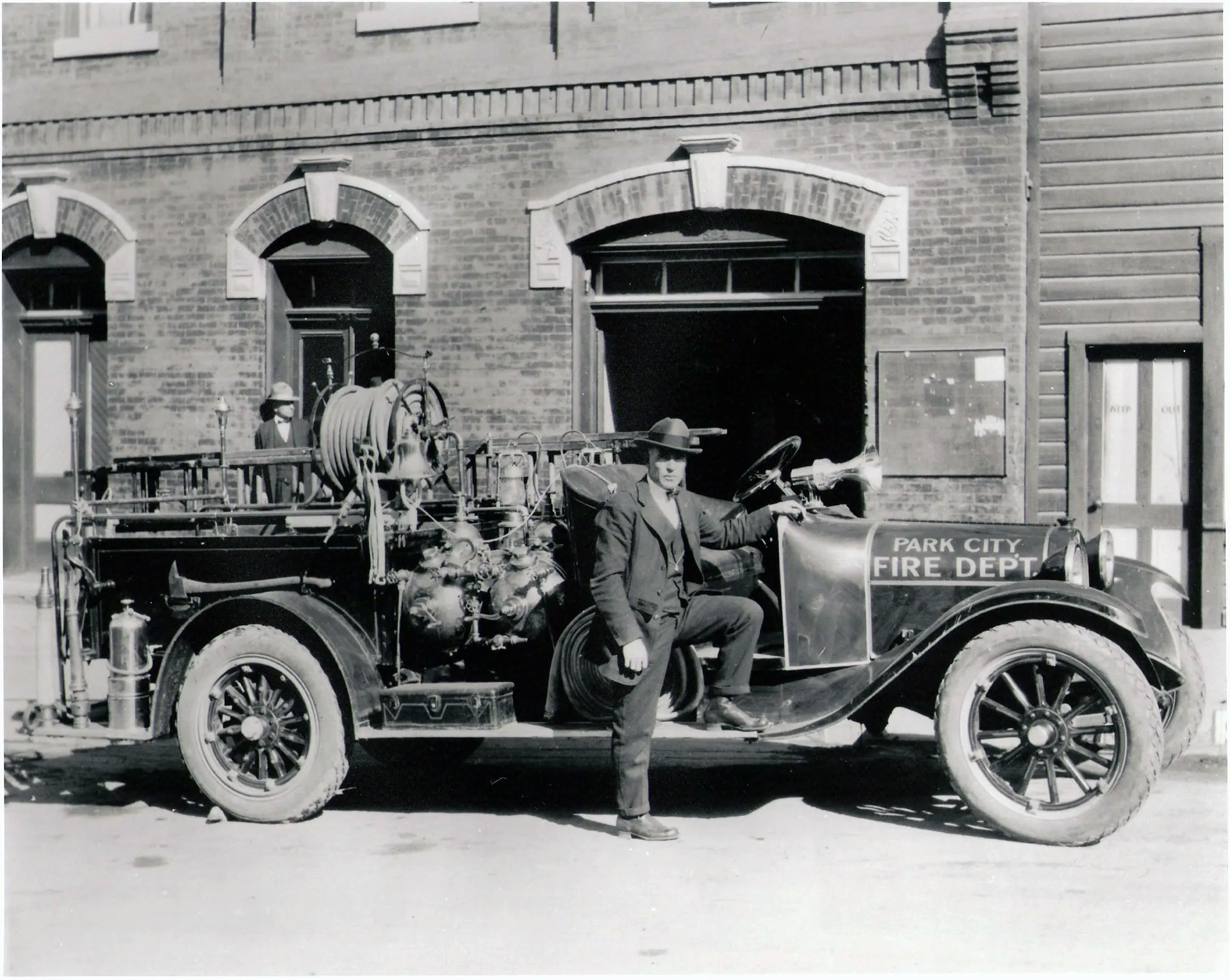 A vintage fire truck parked outside a brick building, with a man in a suit and hat posing next to it, showcasing historical firefighting.