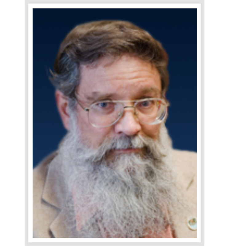The image shows a man with a long beard and glasses, wearing a light-colored jacket, against a dark background.