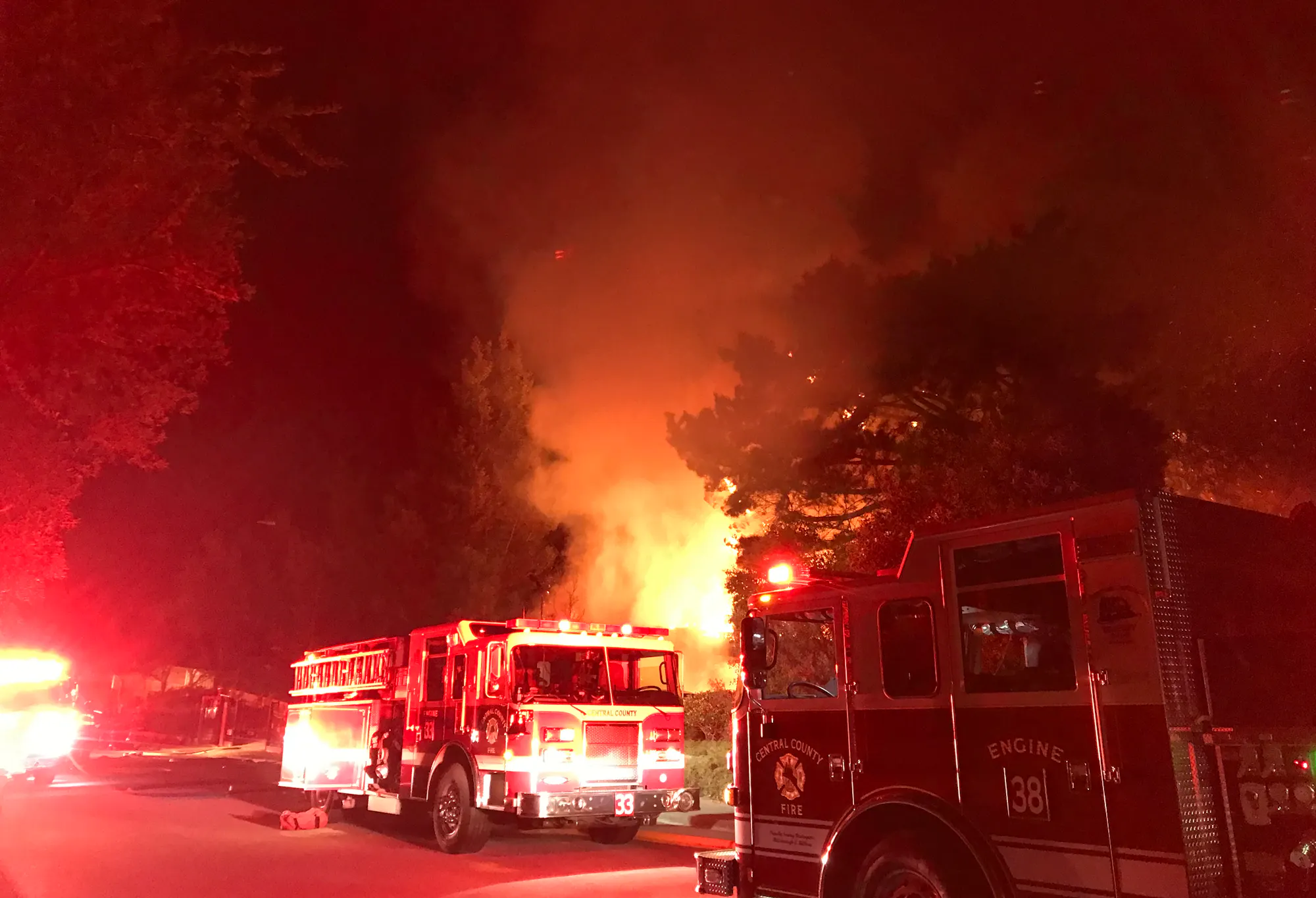 Fire trucks at night responding to a large blaze with smoke and flames visible among trees.