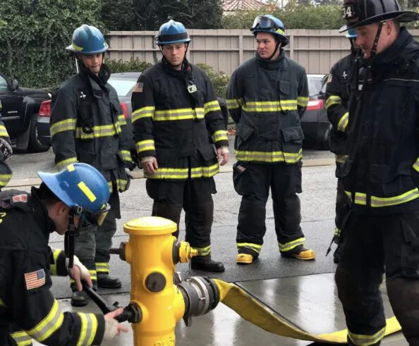 Firefighters in uniform gather around a yellow fire hydrant during a training exercise.