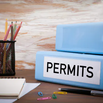The image features two blue folders labeled "PERMITS," a notepad, colored pencils, and paper clips on a wooden desk.
