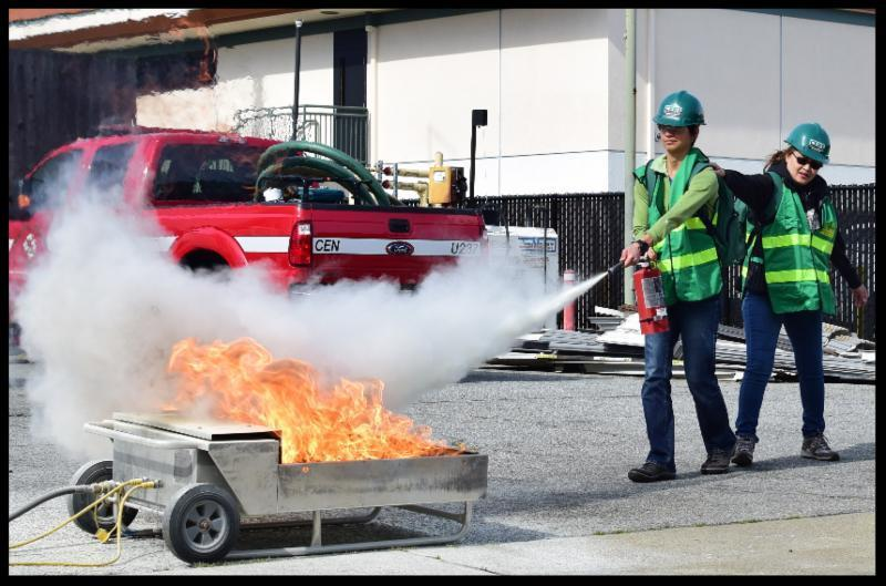 Two people in safety gear extinguish a fire with an extinguisher during a drill.