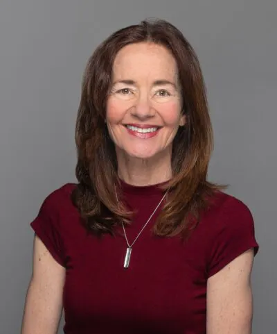 A person with long brown hair, wearing a maroon shirt and a necklace, smiles against a plain gray background.