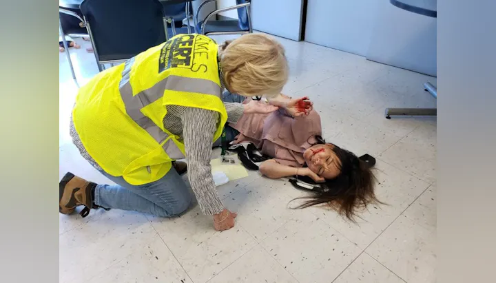 A person in a yellow vest assists a woman lying on the floor with fake injuries, likely during a CERT training exercise.