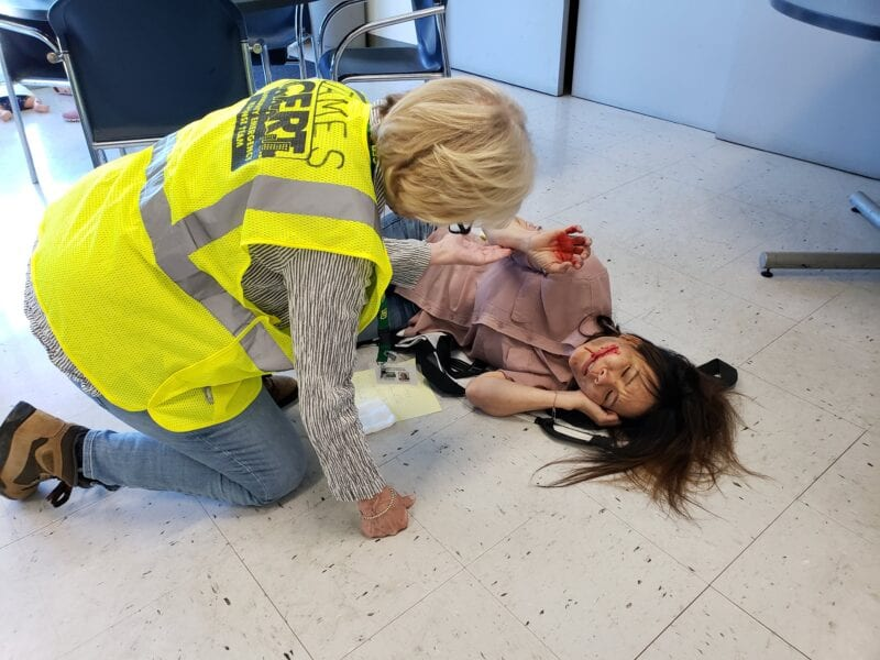 A person in a yellow vest assists a woman lying on the floor with fake injuries, likely during a CERT training exercise.
