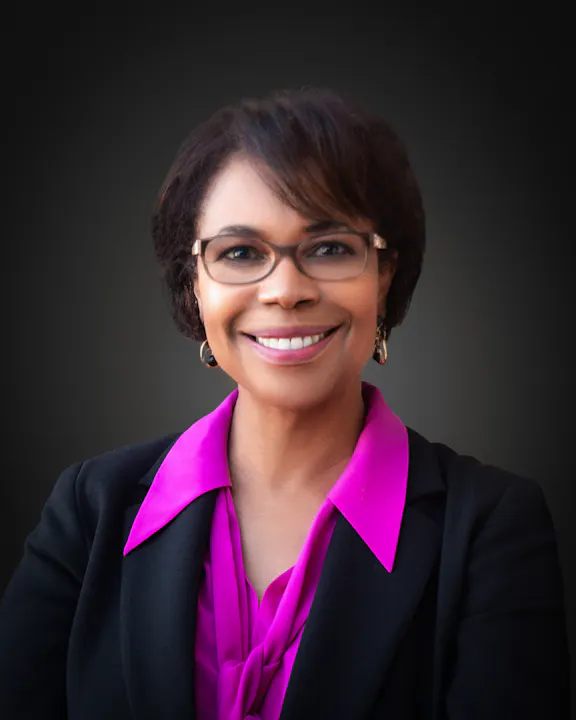 Smiling person wearing glasses, a black blazer, and a bright pink shirt against a dark background.