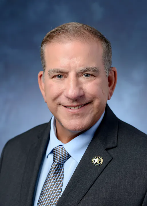 A man in a suit and tie with a badge pin, smiling against a blue-gray background.