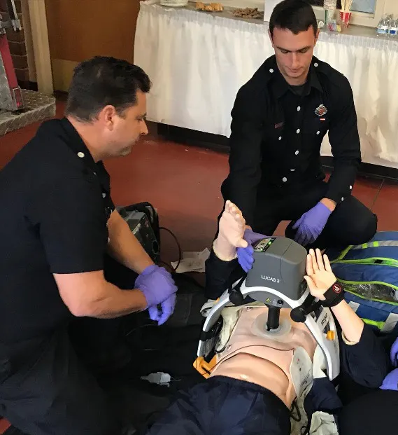Three people in uniform practice CPR on a dummy using a mechanical chest compression device.