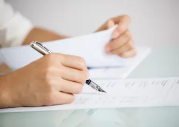 A person is holding a pen and writing on a document while holding another paper in hand.