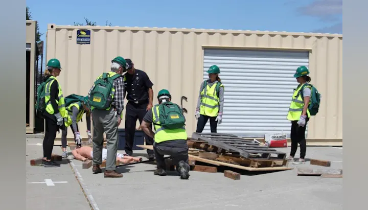 People in safety gear conduct an emergency response drill with a mannequin outside a storage unit.