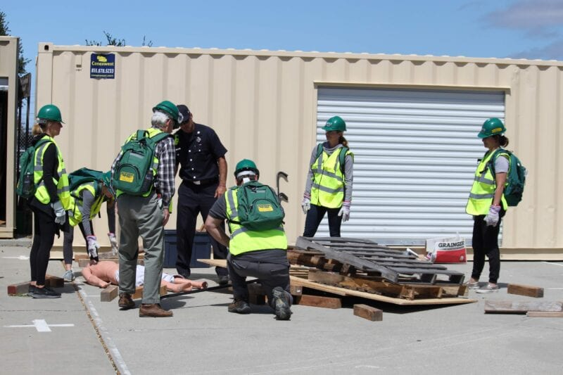 People in safety gear conduct an emergency response drill with a mannequin outside a storage unit.