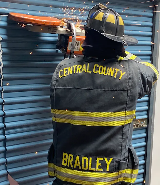 A firefighter from Central County uses a tool to cut through a metal door, creating sparks. The name "Bradley" is visible.