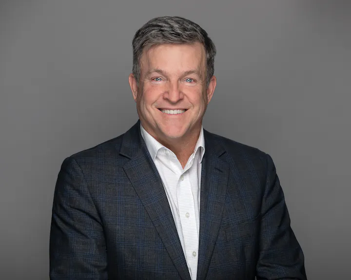 A smiling man in a plaid suit jacket and white shirt against a gray background.