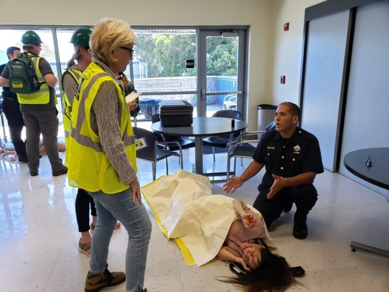 Emergency drill: A responder kneels next to a person covered with a sheet, while others in safety vests observe in a room.