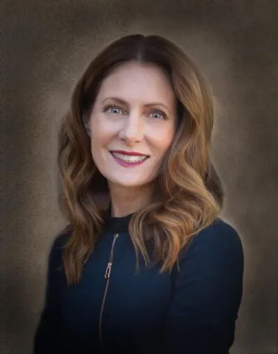 A person with long, wavy brown hair and a black top is smiling against a textured backdrop.