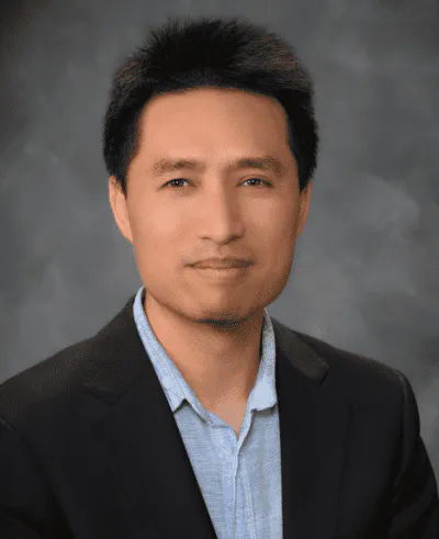 A man in a black suit and light blue shirt, posing in front of a gray background.