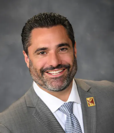 A person in a gray suit and checkered tie, smiling with a pin on the lapel, set against a gray background.