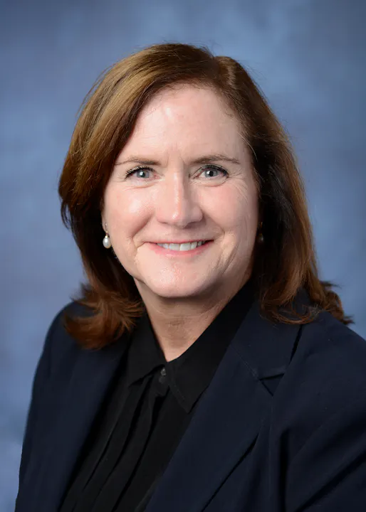 A person with shoulder-length brown hair, wearing a black blazer, in front of a blue background.