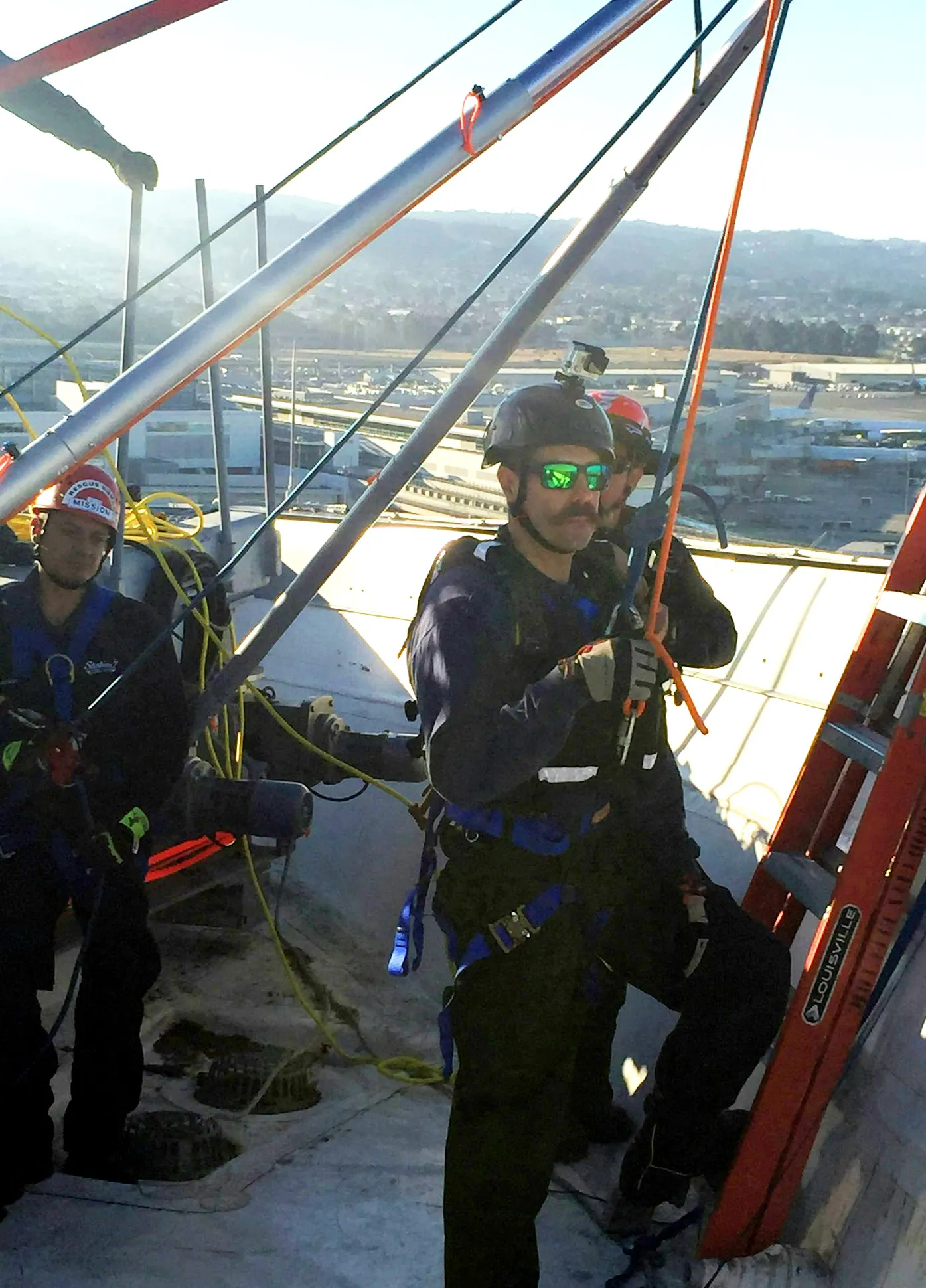 Workers in safety gear on a rooftop, using ropes and ladders, with a cityscape in the background.