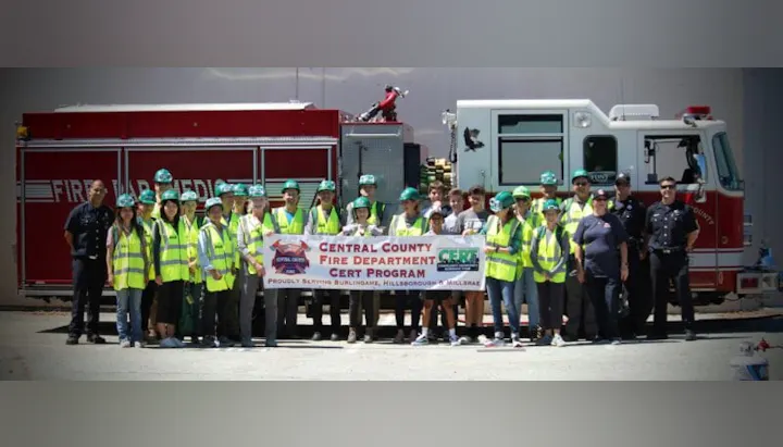 A group of people in reflective vests and helmets stand in front of fire trucks holding a "Central County Fire Department CERT Program" banner.