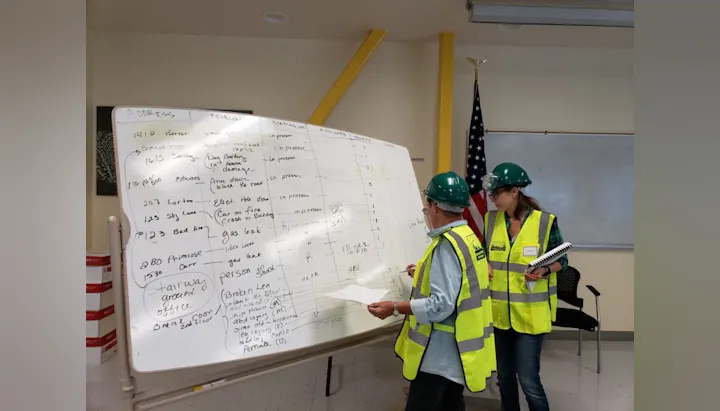 Two people in safety vests and helmets review a whiteboard filled with handwritten notes in a room with an American flag.