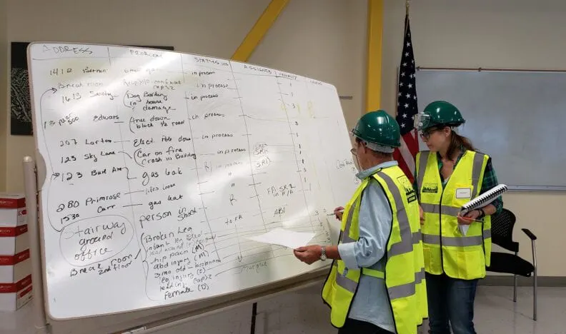 Two people in safety vests and helmets review a whiteboard filled with handwritten notes in a room with an American flag.