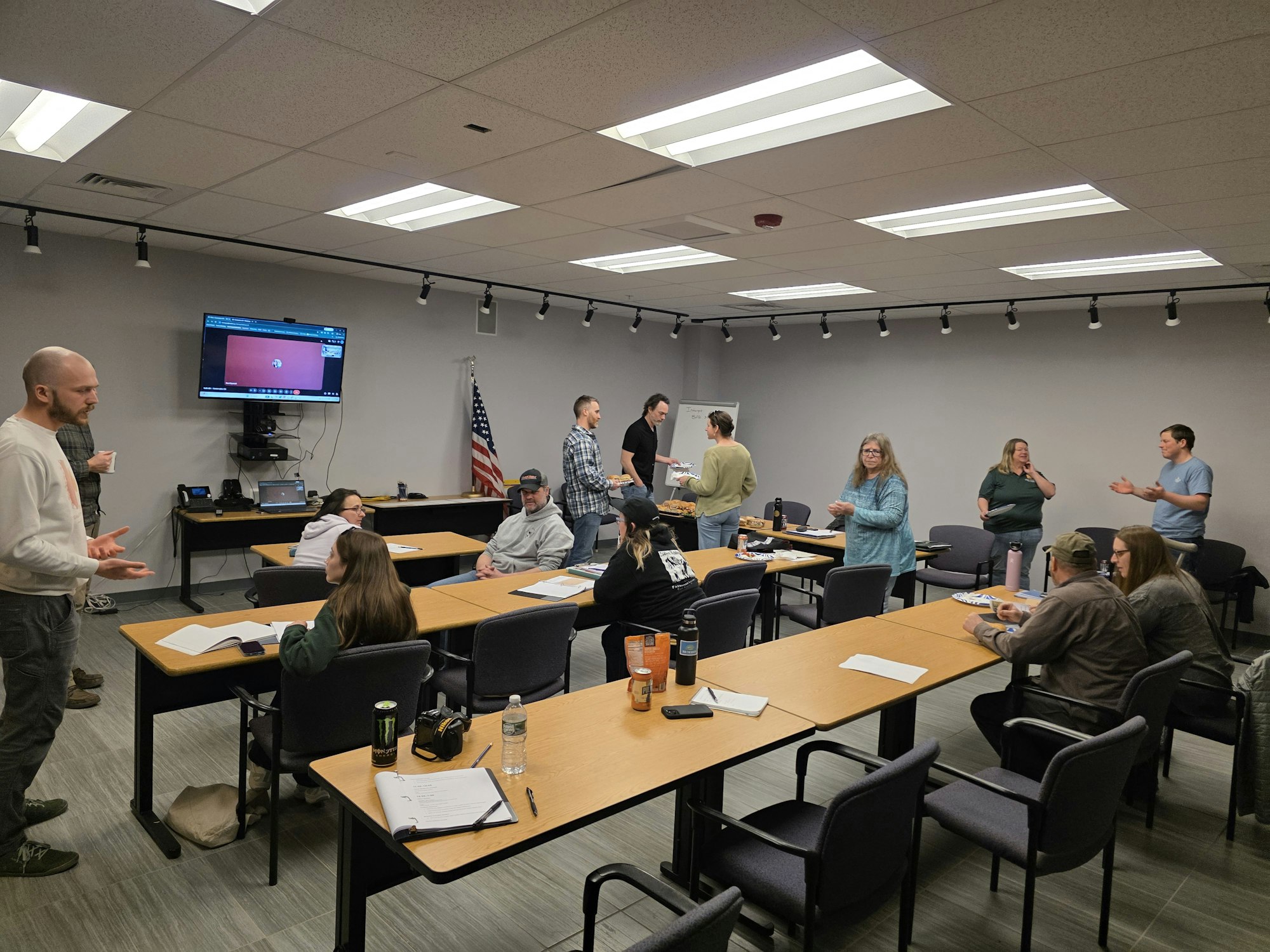 A group of people engaged in discussion and activities in a classroom setting, with tables, chairs, and a presentation screen.