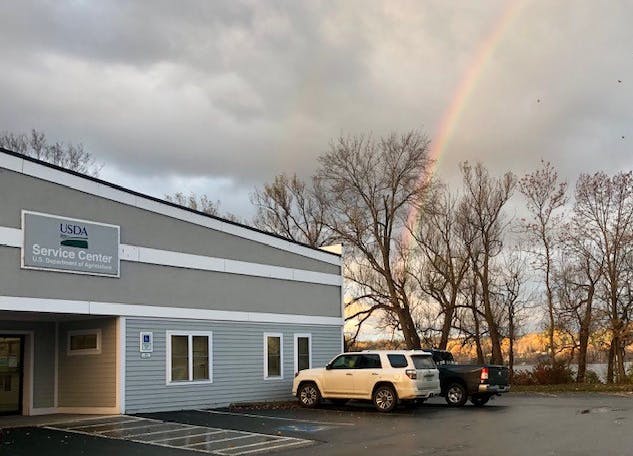 A USDA Service Center building with parked cars, bare trees, and a visible rainbow in the background.