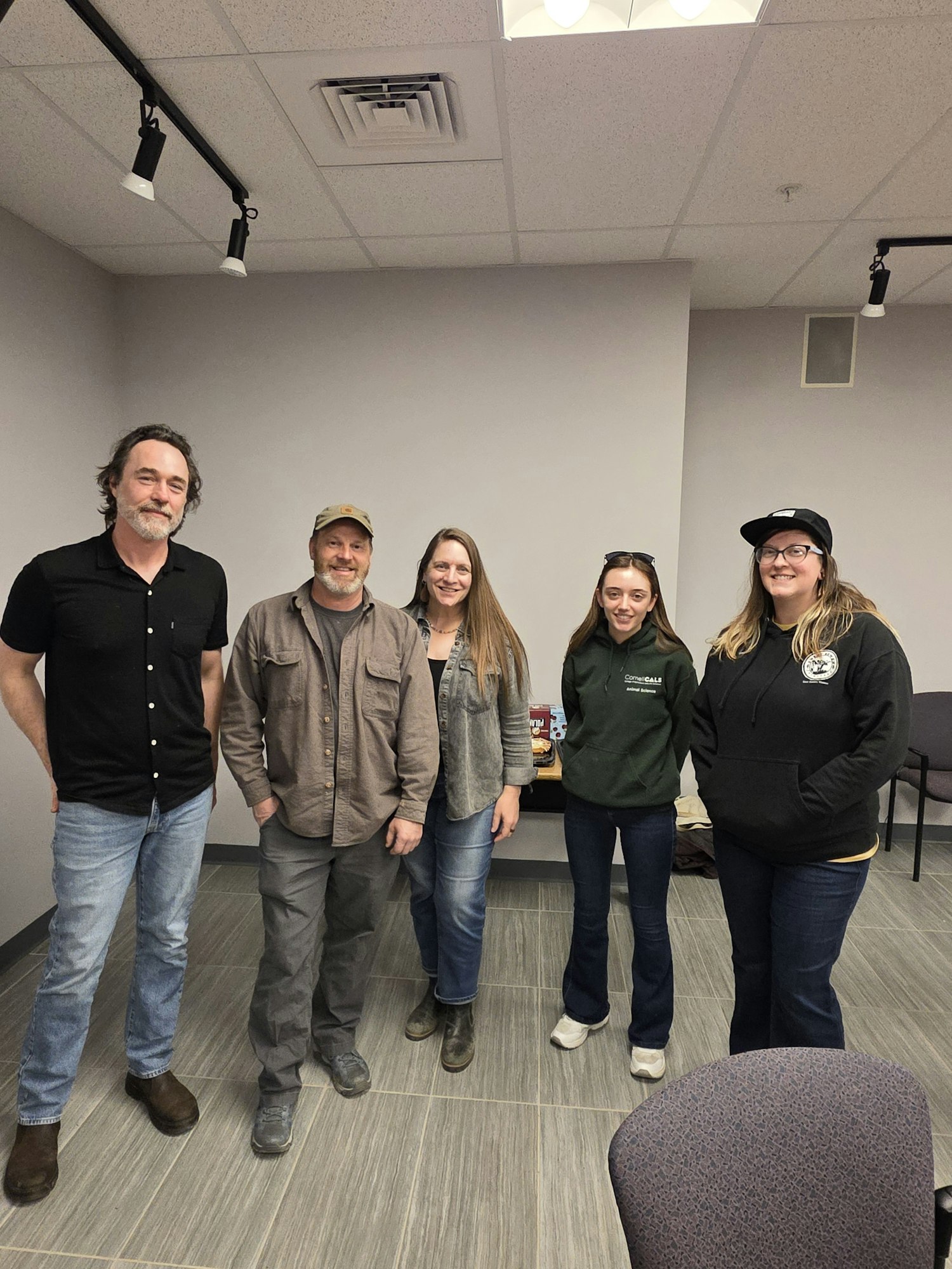 A group of five people poses together in a casual indoor setting, smiling and dressed in relaxed clothing.