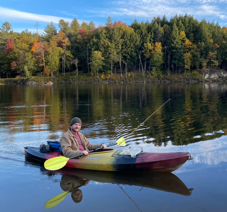 Person fishing from a kayak on a calm lake with a forested shoreline and autumn foliage in the background.