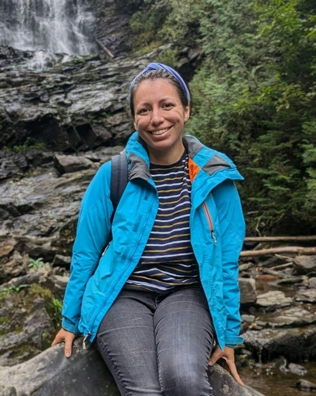 A smiling person seated in front of a waterfall, wearing a blue jacket and headband, surrounded by nature.