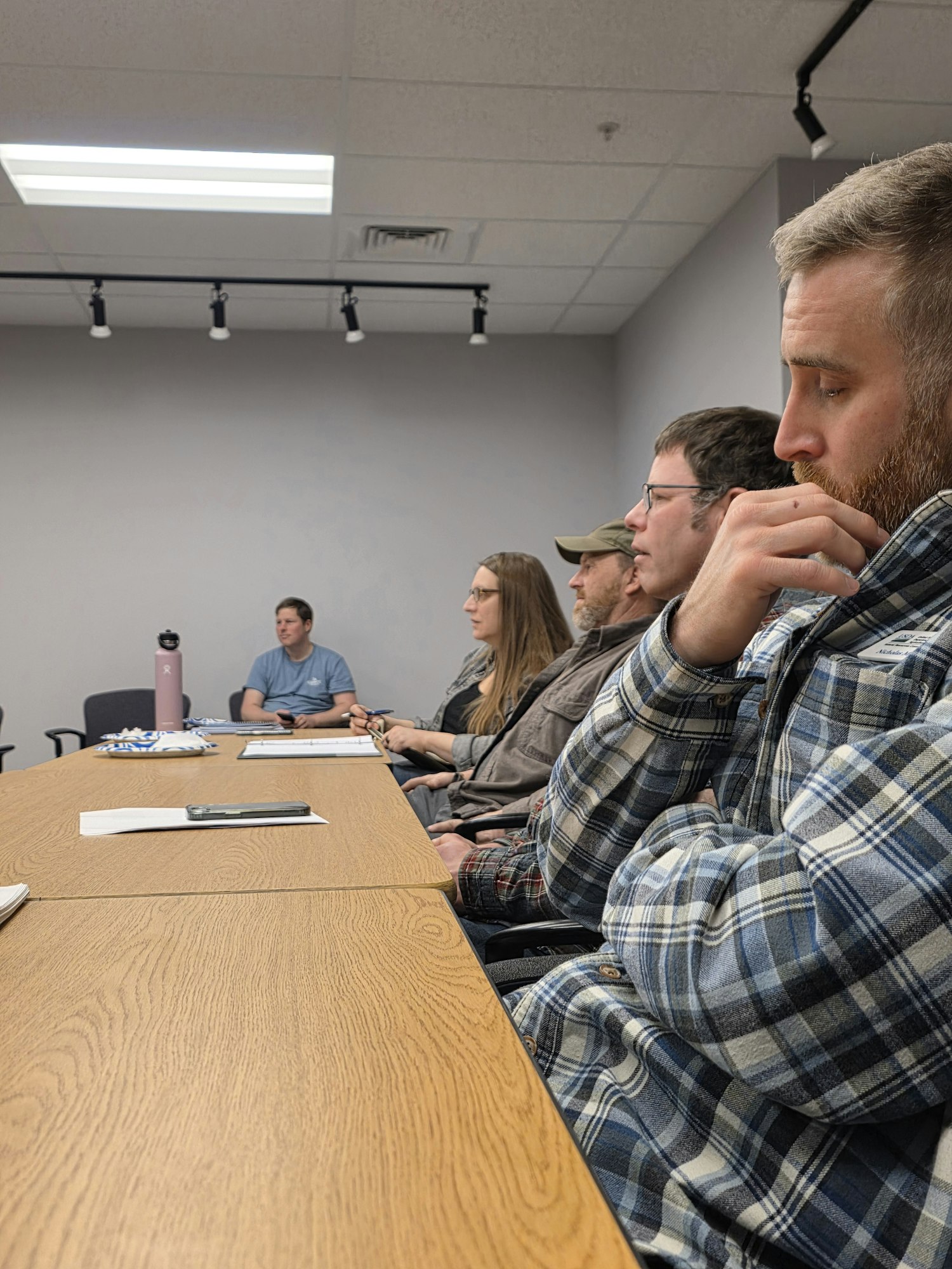 A group of people sitting around a table, appearing focused or engaged, with a mix of casual attire and a gray meeting room.
