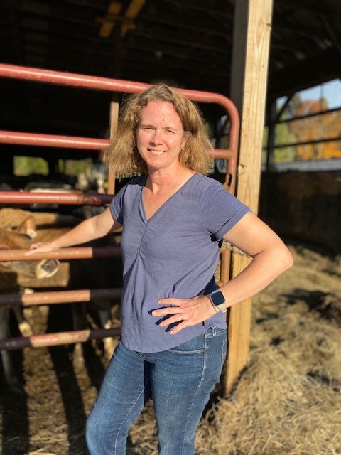 A woman standing by a fence with sunlight behind her, possibly on a farm.