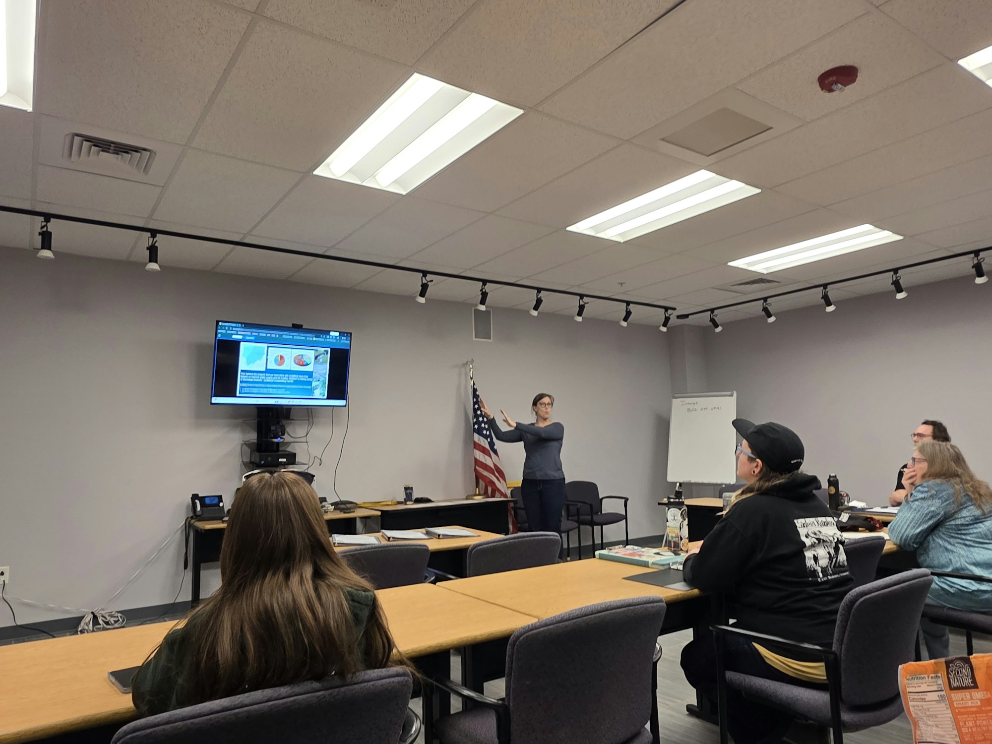 A group in a classroom setting listens to a presentation, with a screen displaying images and information, and an American flag nearby.