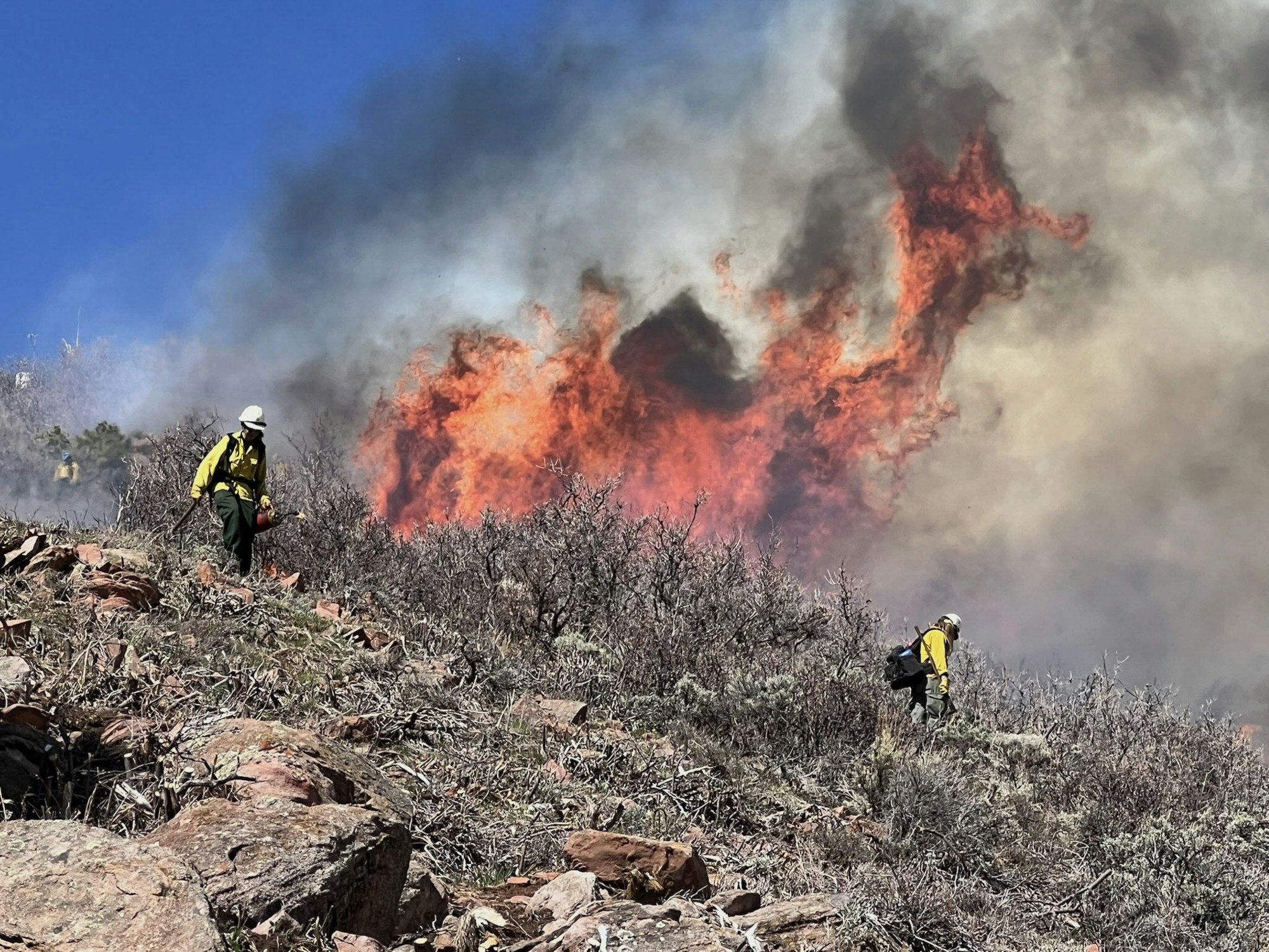 Firefighters work on a hillside with intense flames and smoke in the background.