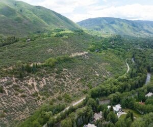 Aerial view of green hills with a river winding through, showcasing a natural landscape. Trees and some homes are visible.