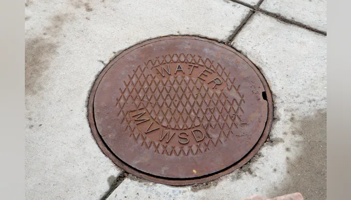 A round, rusty manhole cover labeled "WATER MVWSD" set into concrete pavement.